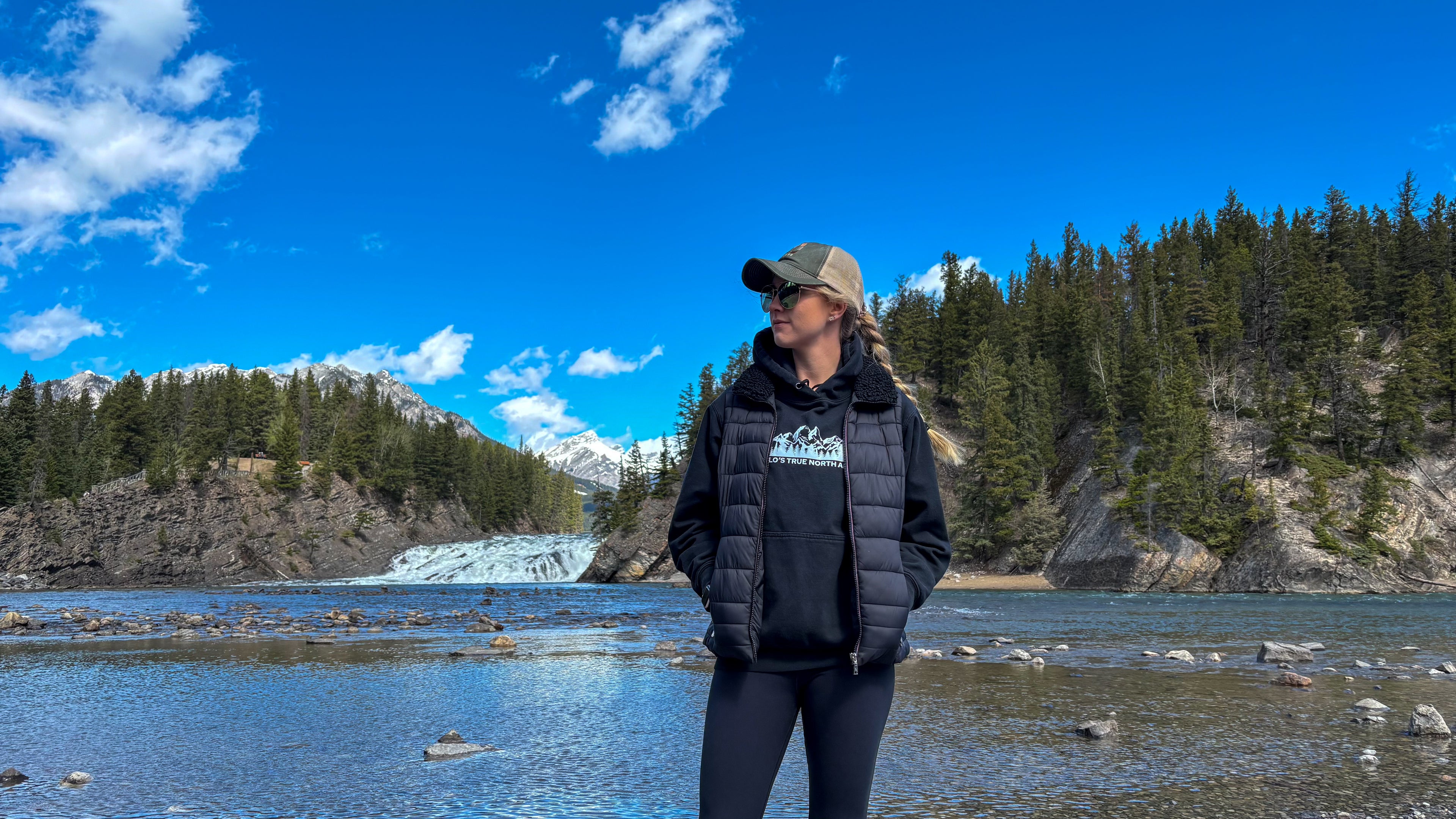 Person standing by a lake with trees and mountains in the background