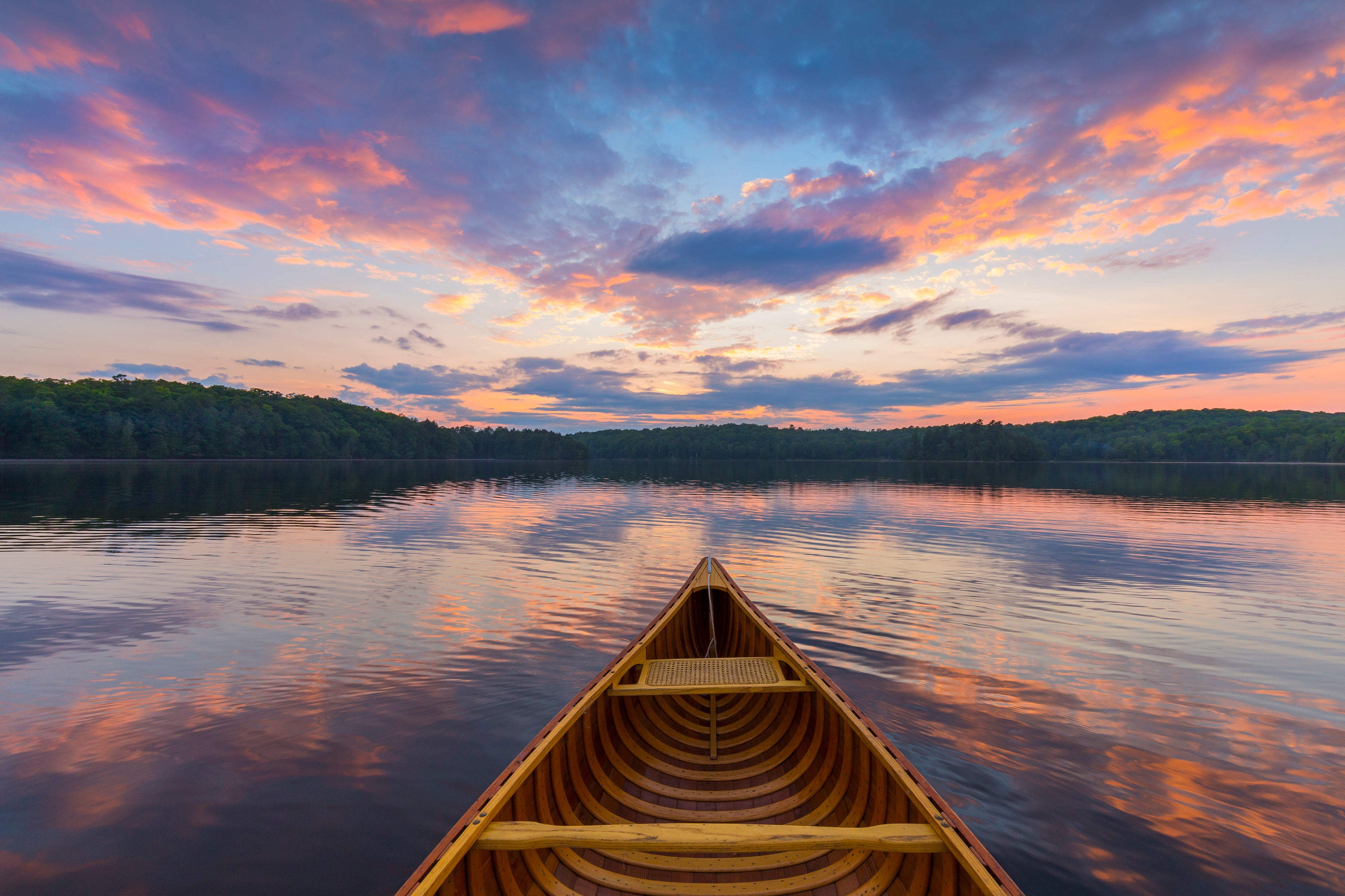 A wooden canoe viewed from the bow, floating on calm lake water at sunset with colorful clouds reflected on the surface and forested shores in the distance.